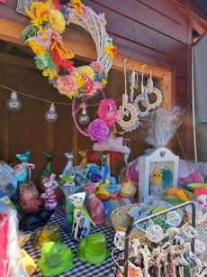 Powiększ zdjęcie: Kolorowe rękodzieło seniorów zaprezentowane na stoisku. Wianki z papieroplastyki. Ceramiczne koty, sowy i żaby.