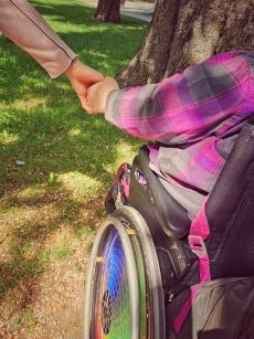 Powiększ zdjęcie: Osoba poruszająca się na wózku inwalidzkim trzyma za rękę z asystenta i idą razem na spacer
