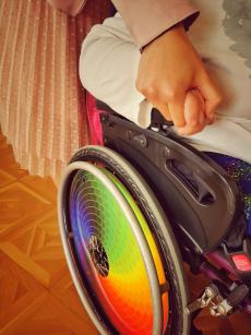 Powiększ zdjęcie: Osoba z niepełnosprawnością siedzi na wózku inwalidzkim i trzyma za rękę kobietę, która jest Asystentem osobistym osoby z niepełnosprawnością. Na zdjęciu widoczne jest również kolorowe, tęczowe koło od wózka inwalidzkiego.