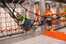 Powiększ zdjęcie: Park trampolin. Zdjęcie przedstawia chłopca, który pokonuje przeszkodę – konstrukcję wykonaną z lin, drugi chłopiec przygląda się temu.  Chłopcy są w parku trampolin.