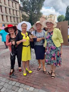 Powiększ zdjęcie: Seniorki w odświętnych kreacjach i własnoręcznie przygotowanych kapeluszach przed Ratuszem