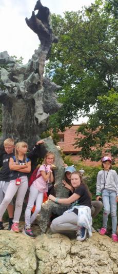 Powiększ zdjęcie: Smok Wawelski. Zdjęcie przedstawia szóstkę dzieci, która pozuje do zdjęcia na tle smoka wawelskiego. Na drugim planie widać drzewo oraz mury Zamku Wawelskiego. 