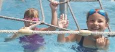 Powiększ zdjęcie: Zabawy w basenie. Zdjęcie przedstawia dwie uśmiechnięte dziewczynki bawiące się w basenie. Dziewczynki trzymają się konstrukcji wykonanej ze sznura.