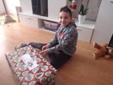 Powiększ zdjęcie: Zdjęcie przedstawia ciemnowłosego, uśmiechniętego chłopca, który siedząc na podłodze rozpakowuje świąteczny prezent.