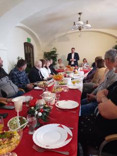Powiększ zdjęcie: Zdjęcie przedstawia grupę ludzi siedzących po obu stronach świątecznie przystrojonego stołu oraz stojącego i przemawiającego Prezydenta miasta Pana Klimaszewskiego.