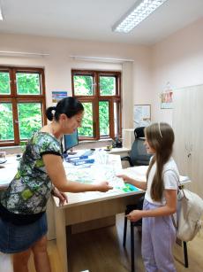 Powiększ zdjęcie: Zdjęcie przedstawia kobietę wręczającą dziewczynce dyplom i nagrodę za udział w konkursie.