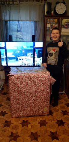 Powiększ zdjęcie: Zdjęcie przedstawia ogromne pudełko – prezent i stojącego obok zadowolonego chłopca na tle okna i włączonego telewizora.