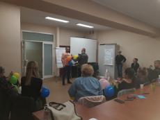 Powiększ zdjęcie: Zdjęcie przedstawia osoby uczestniczące w szkoleniu dla kandydatów na rodziny zastępcze niezawodowe.  Na pierwszym planie widać uczestników szkolenia siedzących w kręgu. Natomiast w głębi stoją trzy osoby, które wykonują ćwiczenie zalecone przez trenera.