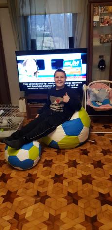 Powiększ zdjęcie: Zdjęcie przedstawia radosnego chłopca na tle okna i włączonego telewizora, który siedzi na pufach w kształcie piłek.