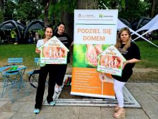 Powiększ zdjęcie: Zdjęcie przedstawia trzy kobiety stojące na tle banera w kolorach zielono pomarańczowym. Dwie z nich przekazują sobie ulotki z informacjami na temat pieczy zastępczej.