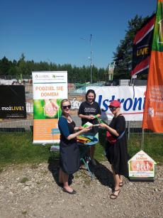 Powiększ zdjęcie: Zdjęcie przedstawia trzy uśmiechnięte kobiety trzymające w dłoniach pomarańczowo-zielone reklamy w kształcie domków z hasłem kampanii Podziel się domem.