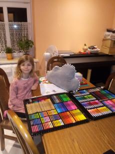 Powiększ zdjęcie: Zdjęcie przedstawia uśmiechniętą dziewczynkę siedzącą na fotelu, przy biurku z dumą prezentując prezent profesjonalny zestaw plastyczny