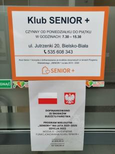 Powiększ zdjęcie: Zdjęcie przedstawia drzwi wejściowe do Klubu Senior+ przy ulicy Jutrzenki 20 w Bielsku-Białej. Na górze drzwi znajduje się plakat z białym tłem i pomarańczową ramką. Granatowymi literami jest napisane: Klub Senior+ czynny od poniedziałku do piątku w godzinach 7.30-15.30, ul. Jutrzenki 20, 43-300 Bielsko-Biała, telefon 535 608 343. Klub Senior+ korzysta z dofinansowania ze środków otrzymanych w ramach Programu Wieloletniego „Senior+” na lata 2015-2020. Na dole jest logo Programu Senior+ czyli pomarańczowy domek z sercem. Poniżej na drzwiach zamieszczono Plakat Informacyjny Programu SENIOR+, który przedstawia u góry flagę i godło Polski, poniżej napis: Dofinansowano ze środków budżetu państwa. Program Wieloletni „Senior+” na lata 2021-2025 edycja 2024. Moduł II – Zapewnienie funkcjonowania Klubu Senior+. Na dole plakatu napis: dofinansowanie 61 920,00 zł całkowita wartość 141 920,00 zł.