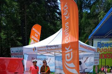 1.	Zdjęcie przedstawia dwoje kobiety stojące pod namiotem obok  pomarańczowo zielonego plakat z napisem podziel się domem. Jedna kobieta w różowej bluzce trzyma rower.