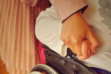 Osoba z niepełnosprawnością siedzi na wózku inwalidzkim i trzyma za rękę kobietę, która jest Asystentem osobistym osoby z niepełnosprawnością. Na zdjęciu widoczne jest również kolorowe, tęczowe koło od wózka inwalidzkiego.