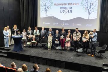 W tle wyświetlony kolorowy napis PRZYJACIELE PROJEKTU PODZIEL SIĘ DOMEM, po bokach drzewa z ptaszkami na gałązkach. Grupa kobiet i mężczyzn oraz dzieci trzymających puzzle i dyplomy. Wszyscy zadowoleni, uśmiechnięci stoją na scenie. Kobieta mówi przez mikrofon.