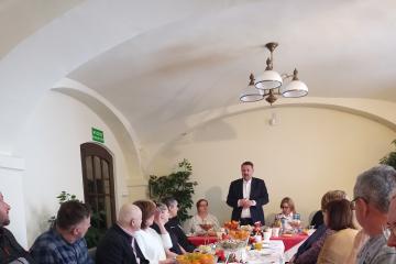Zdjęcie przedstawia grupę ludzi siedzących po obu stronach świątecznie przystrojonego stołu oraz stojącego i przemawiającego Prezydenta miasta Pana Klimaszewskiego.