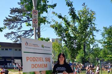 Zdjęcie przedstawia kobietę stojącą obok zielono pomarańczowego plakatu Podziel się domem, który promuje rodzicielstwo zastępcze.